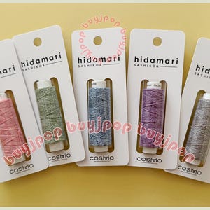 Può includere: Cinque rocchetti di filo colorato su cartoncini bianchi. Ogni cartoncino riporta la scritta "hidamari SASHIKO" e "cosmo". I colori dei fili includono rosa, verde, blu, viola e grigio. I rocchetti sono lunghi circa 2.5 cm.