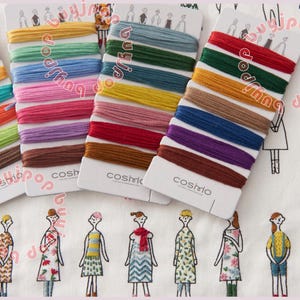 May include: Four sets of Cosmo embroidery floss in various colors, including yellow, green, blue, pink, and brown. The floss is arranged on white cards with the Cosmo logo. The background features a fabric with a repeating pattern of stylized figures.