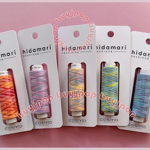 Puede incluir: Cinco carretes de hilo de bordar de colores, cada uno con una etiqueta que dice "hidamari sashiko" y "cosmo". El hilo es en tonos de rosa, amarillo, azul y naranja.