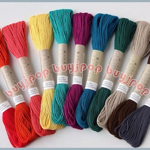 May include: A collection of colorful embroidery floss in shades of red, orange, yellow, teal, purple, blue, green, beige, brown, and navy. Each skein is labeled with Japanese text and a cotton content percentage.