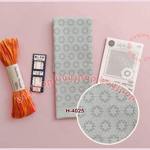 May include: A collection of Sashiko embroidery supplies, including a bundle of orange and yellow thread, needles, a gray fabric piece with a star pattern, and a pattern guide. The fabric is marked with "H-4025".