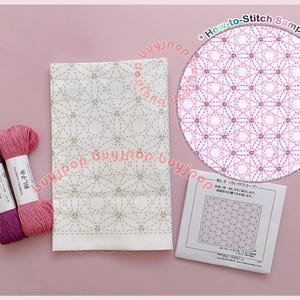 May include: A craft kit with a white fabric panel featuring a geometric stitched design, two spools of thread in pink and purple, and a sample stitch guide. The text "How-to-Stitch Sample" is visible.