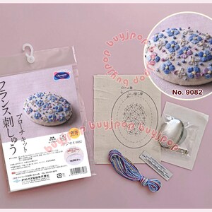 May include: An embroidery kit featuring a finished oval brooch with blue and purple floral stitching, a printed fabric pattern, a metal brooch pin, and a bundle of multi-colored embroidery thread, all packaged with instructional text.