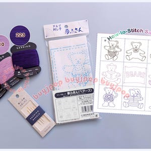 May include: A Sashiko embroidery kit featuring three spools of thread in brown, light purple, and dark purple, a pack of needles, a pre-printed fabric panel with a bear design, and a printed How-to-Stitch Sample sheet showing nine bear illustrations.
