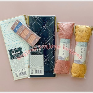 Puede incluir: Una colección de suministros de bordado japonés, que incluye tela, agujas e hilo. La tela es blanca con costuras azules y azul marino con costuras blancas. También se incluyen dos bobinas de hilo rosa y dos bobinas de hilo amarillo.