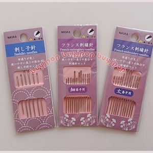 May include: Three packages of embroidery needles are displayed. The packages are lavender with pink inserts holding the needles. Text on the packages includes "NASKA", "Sashiko needles", and "French embroidery needles". The needles are silver.