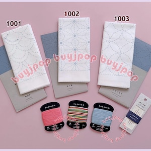 May include: An assortment of Sashiko embroidery supplies, including white fabric with stitched patterns, gray paper, spools of colorful thread, and needles. The fabric designs include geometric and floral patterns. The items are arranged on a pink surface.