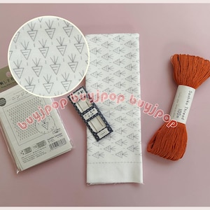 Könnte beinhalten: Eine Sammlung von Nähutensilien auf einer rosa Oberfläche. Enthält einen weißen Stoff mit Karottenmuster, eine Packung Nadeln, eine Packung Nähnadeln und eine Spule orangefarbenen Sashiko-Garns.