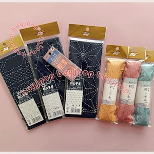 May include: An assortment of Japanese sashiko embroidery supplies. The image shows several packages of navy blue fabric with white stitching, spools of colorful thread, and a package of needles. The packaging has Japanese text.