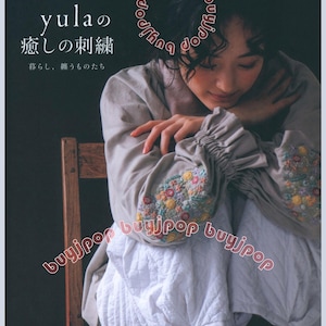 May include: A book cover featuring a woman in a light-colored outfit with embroidered sleeves. The title "yula" and Japanese text are visible. The sleeves are adorned with colorful floral embroidery. The woman is seated on a wooden chair.