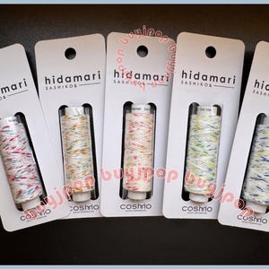 May include: Five spools of Cosmo brand thread, each on a white card with the "hidamari SASHIKO&" logo. The thread colors vary, including red, white, blue, yellow, and orange, creating a speckled effect. The cards have a hole at the top for hanging.