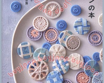Puede incluir: Una colección de botones de hilo hechos a mano en varios colores y patrones, incluyendo azul, blanco y melocotón, dispuestos sobre un plato gris claro. La imagen también presenta texto en japonés.