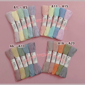 May include: A collection of embroidery floss in various pastel colors, including pink, yellow, green, and blue. Each bundle is labeled with a code, such as A1-A5, A6-A10, A11-A15, and A16-A20. The floss is neatly arranged on a pink background.