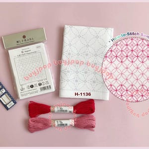 Könnte beinhalten: Eine Sammlung von Sashiko-Stickzubehör, darunter ein weißer Stoff mit einem gestickten Muster, rote und rosa Garnrollen, Nadeln und eine Musteranleitung. Eine Probe der Stickerei ist in einem Kreis dargestellt.