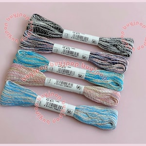 May include: Five skeins of embroidery floss in various colors, including black, purple, teal, pink, and blue. Each skein has a white label with black text and a barcode. The floss is neatly arranged on a light pink background.