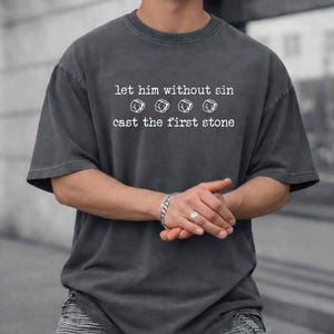 May include: A charcoal gray short-sleeved t-shirt featuring white text that reads "let him without sin" followed by four small stone illustrations and the text "cast the first stone" in a typewriter-style font across the chest area.