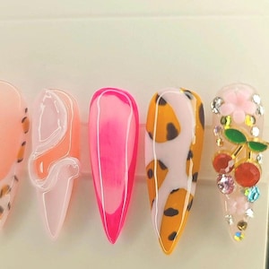 May include: Five long, pointed false nails with different designs. The first nail is peach with a leopard print. The second nail is white with a swirl design. The third nail is pink with a white line down the centre. The fourth nail is white with a leopard print in orange and brown. The fifth nail is white with a cherry design and rhinestones.