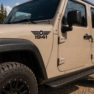 May include: A tan Jeep SUV with black tires and a black side step. A black graphic with wings, a star, and the number 1941 is on the front panel of the vehicle.