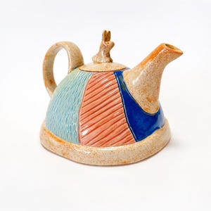 Puede incluir: Una tetera de cerámica con un diseño caprichoso. El cuerpo presenta secciones en azul, coral y azul, con una superficie texturizada. El pico y el asa son de color beige, y una pequeña figura de conejo adorna la tapa. Una pieza única para la decoración de la cocina.