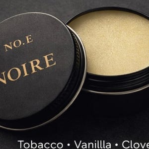 Op de afbeelding: Een zwarte container met een deksel, met de gouden letters "NO.E NOIRE". De container is open en onthult een lichtgekleurde balsem. De onderkant van de afbeelding vermeldt "Tobacco • Vanilla • Clove".