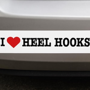 Può includere: Un adesivo rettangolare bianco con la scritta "I ❤️ HEEL HOOKS" in nero, con un simbolo a cuore rosso. L'adesivo è attaccato alla parte posteriore di un veicolo.
