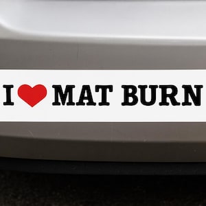 Könnte beinhalten: Weißer rechteckiger Aufkleber mit dem Text "I ❤️ MAT BURN" in Schwarz und einem roten Herzsymbol. Der Aufkleber ist an der Rückseite eines Fahrzeugs angebracht.