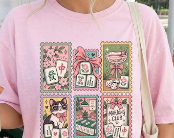 May include: A light pink t-shirt featuring a graphic print of six rectangular postage stamp designs. The illustrations include mahjong tiles, a cat wearing glasses, a cocktail glass, and a sign that reads Mahjong Club, all in pastel colors.