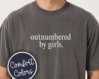 May include: A charcoal grey short-sleeve t-shirt featuring the white text "outnumbered by girls." on the chest. A circular navy blue badge with the white text "Comfort Colors" is positioned on the left side of the shirt over the chest area.