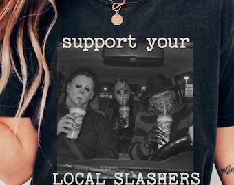 May include: A charcoal gray short-sleeve t-shirt featuring a black and white graphic of three horror movie characters sitting in a car drinking from cups. The text above and below the graphic reads support your LOCAL SLASHERS in white.