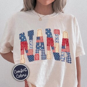 May include: Cream-colored t-shirt with the word "NANA" in a patchwork design featuring red, white, and blue patterns. The shirt is paired with red and white checkered shorts. A circular "Comfort Colors" logo is visible.