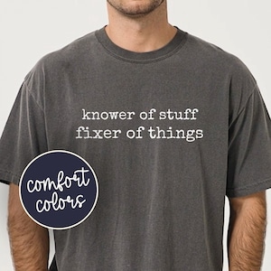 May include: A charcoal gray short-sleeve t-shirt featuring the white text "knower of stuff fixer of things" printed on the chest. A circular navy blue logo with the white text "comfort colors" is positioned on the left side of the shirt.