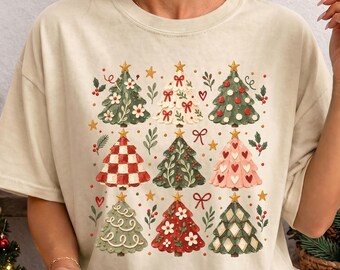 May include: A beige short-sleeve t-shirt featuring a graphic print of nine stylized Christmas trees in various patterns and colors, including red, green, and pink, accented with small stars, hearts, and bows, paired with a patterned mug.