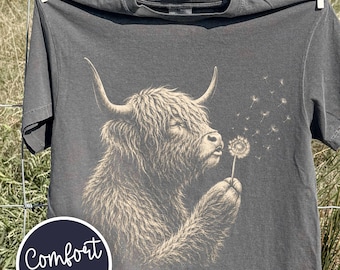 May include: A charcoal gray short-sleeve t-shirt featuring a graphic illustration of a Highland cow blowing on a dandelion. The cow is rendered in light tan lines, and the text Comfort Colors is printed in a white circle on the lower left side.