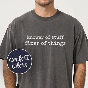 May include: Dark gray t-shirt with the text "knower of stuff fixer of things" in white. A navy blue circle with the words "comfort colors" is on the left side of the shirt. The shirt has short sleeves and a crew neck.