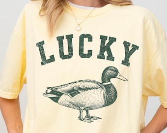 May include: A pale yellow short-sleeve t-shirt featuring the word LUCKY in dark green block letters above a detailed illustration of a mallard duck. The graphic has a distressed, textured appearance, and the shirt has a crew neckline.