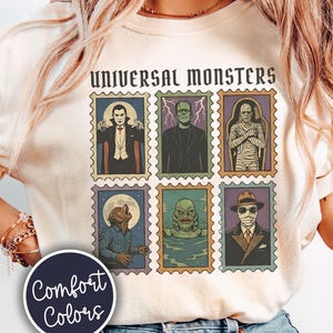 May include: A cream-colored t-shirt featuring a graphic print of six postage stamp-style illustrations of Universal Monsters, including Dracula, Frankenstein, the Mummy, the Wolf Man, the Creature from the Black Lagoon, and the Invisible Man.