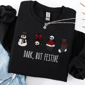 Op de afbeelding: Een zwart sweatshirt met lange mouwen en een afbeelding van een sneeuwpop, maretak met een schedel, een cupcake met een schedel en een kat in een kerstsok. De tekst "DARK, BUT FESTIVE" staat in witte letters. Gecombineerd met een blauwe spijkerbroek.