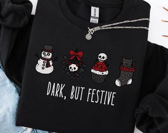 May include: A black long-sleeve sweatshirt featuring a graphic print of a snowman, mistletoe with a skull, a cupcake with a skull topper, and a cat in a stocking. The text "DARK, BUT FESTIVE" is printed in white below the holiday-themed illustrations.