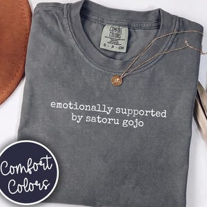 May include: A charcoal grey short-sleeve t-shirt with the white text "emotionally supported by satoru gojo" printed on the chest. The shirt features a Comfort Colors brand label at the inner neckline and a small circular logo sticker.