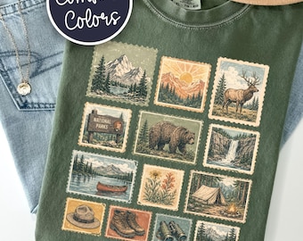 May include: An olive green t-shirt featuring a grid of twelve postage stamp-style illustrations depicting outdoor nature scenes, including mountains, a bear, an elk, a waterfall, a canoe, a tent, hiking boots, binoculars, and a park sign.
