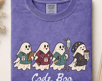 May include: A purple crewneck t-shirt featuring a graphic of four cartoon ghosts dressed as medical staff. The ghosts wear scrubs, a stethoscope, and a hat, with medical equipment and the text Code Boo printed below in a white script font.