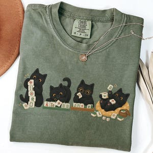 May include: An olive green short-sleeve t-shirt featuring a graphic of four black cats playing with mahjong tiles. The shirt has a crew neckline and a Comfort Colors brand label on the inside collar. The graphic shows the cats stacking, arranging, and lounging with various mahjong tiles, including one cat wearing round glasses.