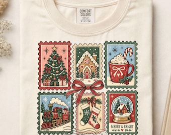May include: A cream-colored t-shirt featuring a graphic print of six vintage-style postage stamps depicting Christmas scenes, including a decorated tree, gingerbread house, cocoa mug, steam train, stockings, and snow globe, tied with a red bow.