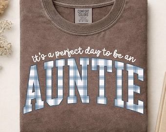 May include: A brown crewneck t-shirt with a vintage wash finish. The front features the white text "it's a perfect day to be an" above the word "AUNTIE" in a large, arched, blue and white plaid block font. The inner neck label reads Comfort Colors.