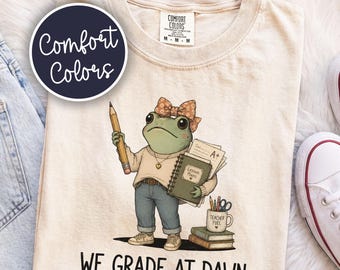 May include: A cream-colored t-shirt featuring a graphic of a frog wearing a bow, a sweater, and jeans. The frog holds a pencil and a notebook labeled Lesson Plans with an A+ paper. Below, the text reads WE GRADE AT DAWN. A mug labeled Teacher Fuel sits on books.