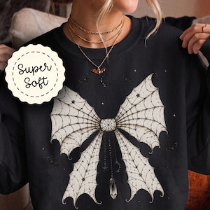 May include: A black long-sleeve sweatshirt featuring a large, off-white bow graphic with a spiderweb pattern. The design includes hanging crystal and moon charms, small stars, and bats. A circular white badge with the text Super Soft is visible.