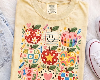 May include: A pale yellow t-shirt featuring a graphic print of nine stylized apples. The apples are decorated with various patterns including daisies, smiley faces, rainbows, hearts, polka dots, and numbers. The design includes small crayons and paperclips.