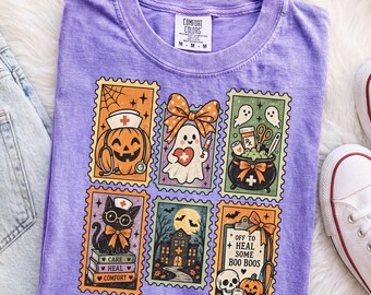 May include: A purple t-shirt featuring a graphic of six rectangular postage stamps with Halloween and medical themes, including a pumpkin with a stethoscope, a ghost with a heart, a cauldron, a cat on books, a haunted house, and a clipboard.