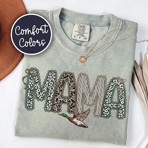 Peut inclure: Un t-shirt vert sauge avec le mot MAMA en grandes lettres aux bords festonn&eacute;s. Les lettres pr&eacute;sentent des motifs l&eacute;opard et ray&eacute;s. Un canard colvert est imprim&eacute; sous le texte. Le t-shirt porte une &eacute;tiquette Comfort Colors au col.