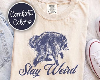 May include: A beige crew neck t-shirt featuring a dark blue graphic of a raccoon and the text "Stay Weird" in a cursive font. The shirt has a Comfort Colors brand tag at the neck, with a size M label visible on the tag.